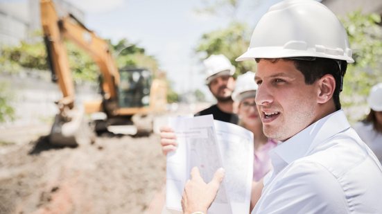 joão campos usa capacete de obras, está com documento em mãos. ao fundo retroescavadeira e um buraco grande