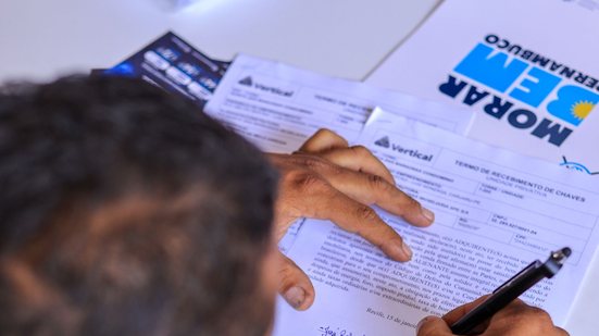 Visão de cima de homem assinando documento. Ao fundo pasta com a logo do Morar bem