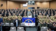 Em auditório, uma fileira de homens estão com farda da PM, acima deles diversas fileiras pessoas usam ternos pretos. No centro está Raquel Lyra segurando bandeira de Pernambuco