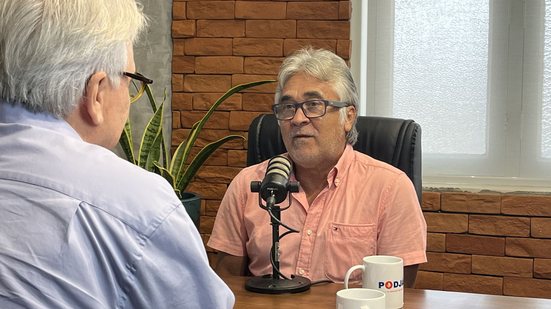 Edmilson Cupertino sentado fala em microfone de podcast. Na mesa uma caneca do PodJá. Em primeiro plano de costas está Jamildo