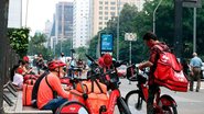 Homens com bolsas térmicas do ifood e rappi parados em praça. Alguns estão de bicicletas