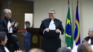 Erik Simões sorri com roupa de magistrado. Ele está ao lado de púlpito. Diversas pessoas em sua frente aplaudem. Ao fundo bandeira do Brasil e de Pernambuco