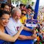Raquel Lyra aponta para bandeira de Pernambuco pequena que Janja segura. Lula e João Campos sorriem. Ao fundo multidão embaixo. Todos usam roupas carnavalescas