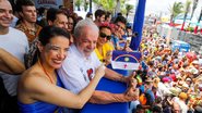 Raquel Lyra aponta para bandeira de Pernambuco pequena que Janja segura. Lula e João Campos sorriem. Ao fundo multidão embaixo. Todos usam roupas carnavalescas