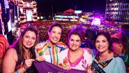 Com roupas carnavalescas, Raquel Lyra, Priscila Krause e duas mulheres sorriem. Ao fundo Palco do Marco Zero com multidão