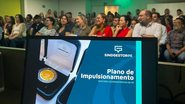 Tela de notebook com imagem escrito "plano de impulsionamento gestores governamentais de pe" e logo do SindGestorPE. Ao fundo pessoas sentadas em auditorio