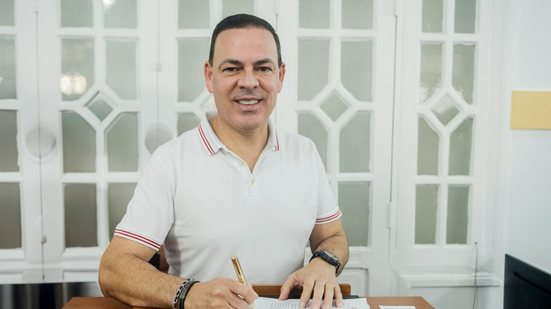 Sivaldo Albino sorri enquanto assina documento, ele está sentado