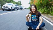 Raquel Lyra sentada no chão de uma estrada sorrindo. Ao fundo carros passam. Ela está com uma camiseta escrito pernambuco e segura uma xicara