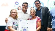 Victor Marques abraçado com mulher loira branca, uma senhora idosa branca e um homem adulto branco de cabelos escuros. Eles seguram documento com imagem escrito "Agora sim a casa é sua"