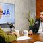 Douglas Nóbrega e Jamildo Melo estão frente a frente sentados em uma mesa em frente a microfones de podcasts. Na mesa canecas do PodJá. Ao fundo, televisão com logo do PodJá na tela