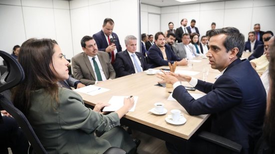 em mesa de reuniao raquel lyra anota em papel enquanto observa fabio aragao que fala ao fundo diversos politicos estao sentados em mesa de reuniao
