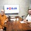 Lula da Fonte e Jamildo estão sentados frente a frente em uma mesa de madeira. Eles estão em frente de microfones de podcast e com xícaras escrito "podjá". Ao fundo, televisão tem "podjá" escrito na tela. Ambos sorriem