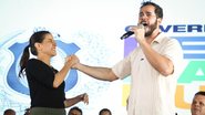 Túlio Gadelha fala com a boca aberta parecendo gritar em um microfone. Raquel sorri enquanto segura sua mão. Ao fundo telão do Governo de Pernambuco