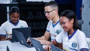 3 jovens, 2 garotas negras e 1 garoto pardo sorriem enquanto olham cada um para um notebook. Eles usam farda do Governo do Estado