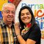 Joselito e Viviane sorriem. Joselito usa roupa xadrez, ela usa um lenço colorido no pescoço com camiseta preta. Ao fundo painel colorido escrito "pernambuco meu paíse são joão"