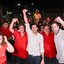 Ao redor de diversas pessoas de vermelho estão Marília Arraes, Lula Cabral, João Campos, Batista Cabral, Pedro Campos e Carlos Costa sorrindo em pé. Lula, João e Pedro estão com os braços erguidos