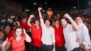 Ao redor de diversas pessoas de vermelho estão Marília Arraes, Lula Cabral, João Campos, Batista Cabral, Pedro Campos e Carlos Costa sorrindo em pé. Lula, João e Pedro estão com os braços erguidos