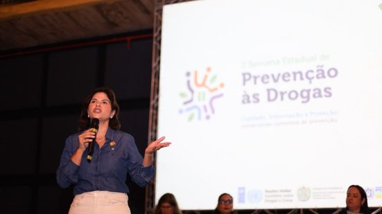 Priscila Krause fala em microfone em frente a um painel escrito prevenção às drogas