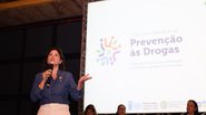 Priscila Krause fala em microfone em frente a um painel escrito prevenção às drogas