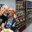 Pollyana Abreu, Raquel Lyra e uma mulher negra sorriem usando toucas na cabeça. Raquel segura junto a mulher um prato de comida. à esquerda dispensa cheia de comida
