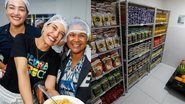 Pollyana Abreu, Raquel Lyra e uma mulher negra sorriem usando toucas na cabeça. Raquel segura junto a mulher um prato de comida. à esquerda dispensa cheia de comida