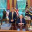 Em salão da Casa Branca com bandeira dos EUA, sorriem Donald Trump e Flávio Bolsonaro. Donald está sentado, Flávio em pé ao seu lado