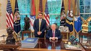 Em salão da Casa Branca com bandeira dos EUA, sorriem Donald Trump e Flávio Bolsonaro. Donald está sentado, Flávio em pé ao seu lado