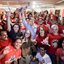 João Campos com as mãos levantadas ao redor de diversas pessoas com camisetas vermelhas. Entre elas Humberto Costa, Rosa Amorim, Ivete Caetano, Kari Santos, Felipe Cury e Dani Portela. Com roupas cinzas estão Marília Arraes e Carlos Costa. Todos sorriem