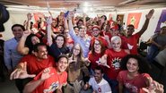 João Campos com as mãos levantadas ao redor de diversas pessoas com camisetas vermelhas. Entre elas Humberto Costa, Rosa Amorim, Ivete Caetano, Kari Santos, Felipe Cury e Dani Portela. Com roupas cinzas estão Marília Arraes e Carlos Costa. Todos sorriem