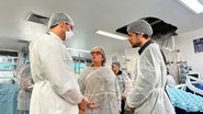 Zilda Cavalcanti e Renato Cirne com toucas e aventais médicos conversam com homem de touca, avental e máscara. Ao fundo, área com teto quebrado e equipamentos médicos