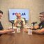 Frente a frente em uma mesa estão Felipe Alecrim e Rinaldo Júnior, que estão em frente a microfones de podcast, no meio na ponta da mesa Jamildo observa os dois. Ao fundo televisão com logo do PodJá