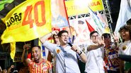 João campos fala em microfone enquanto sorri e usa praguinha no peito. Ao lado está Victor Marques. Diversas bandeiras amarelas escrito João 40 estão ao redor