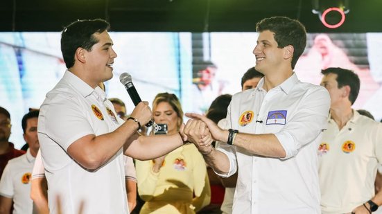 Cayo Albino fala ao microfone, com João Campos no palco