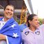 Sorriem lado a lado Fred Loyo e Raquel Lyra. Raquel usa adesivos de coração com o número 45 e acena olhando para o lado. Fred Loyo sorri com uma bandeira enrolada no ombro