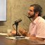 Túlio Gadelha fala em microfone sentado. Ao fundo TV com a logo do PodJá