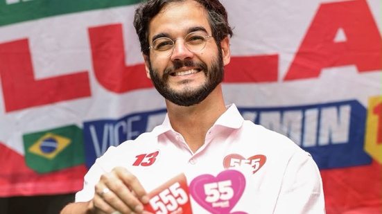 Túlio Gadelha fala com a boca aberta parecendo gritar em um microfone. Raquel sorri enquanto segura sua mão. Ao fundo telão do Governo de Pernambuco