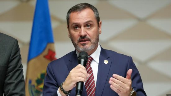 Andrei Mendonça é um homem branco de cabelos e barba grisalha com olhos verdes. Ele usa terno com broche da Polícia Federal enquanto fala em microfone