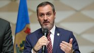 Andrei Mendonça é um homem branco de cabelos e barba grisalha com olhos verdes. Ele usa terno com broche da Polícia Federal enquanto fala em microfone