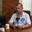 Carlos Sant'anna sentado em mesa de frente para microfone de mesa. ele usa dois adesivos com seu rosto em uma camisa social azul clara