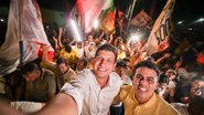 João Campos sorri ao lado de Vinicius Marques, ao fundo diversas pessoas seguram bandeiras. Multidão à noite