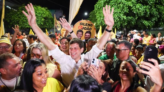 João Campos sorri com braços levantados ao redor de multidão na rua de noite