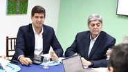 João Campos sentado em mesa ao lado de homem branco idoso. ambos usam ternos. Há outras pessoas fora do foco