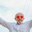 Imagem mostra o presidente Lula em ato político, com camisa branca e óculos vermelho, em formato de estrela, sorrindo e acenando para pessoas