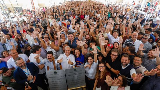 Vista ampla com autoridades e dezenas de moradores acenando para a câmera atrás das placas inaugurais