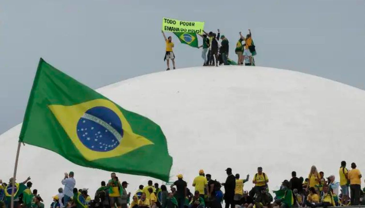 Bandeira do Brasil em primeiro plano, ao fundo vários extremistas praticando atos de vandalismo no DF