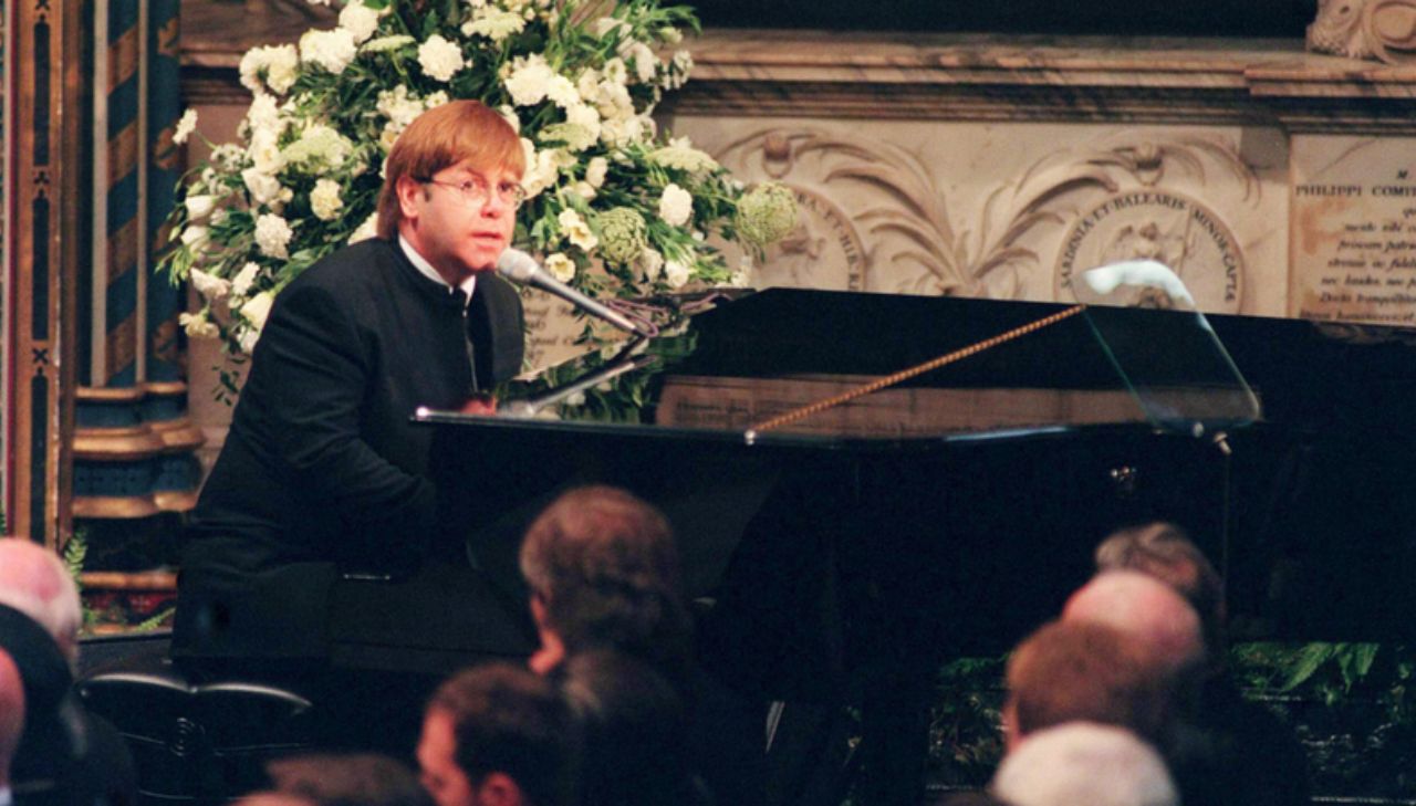 Elton John em um piano, vestido de preto, enquanto toca a música Candle in the Wind no funeral da princesa Diana