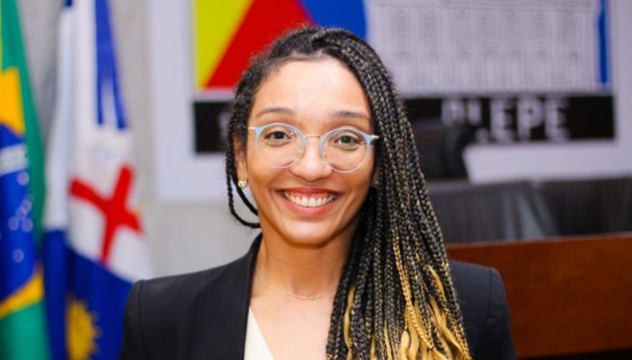 Imagem de uma mulher negra de pele clara, com cabelo trançado preto na raiz e loiro apenas nas pontas. Ela usa óculos e sorri para câmera, ao fundo estão a bandeira do Brasil, de Pernambuco e a marca escrito "Alepe"