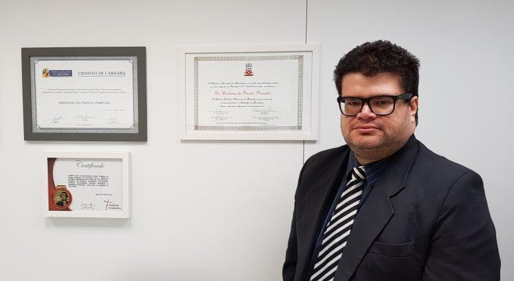 Imagem de um homem branco de cabelo curto pretos com terno e gravata ao lado de diversos certificados em uma parede branca