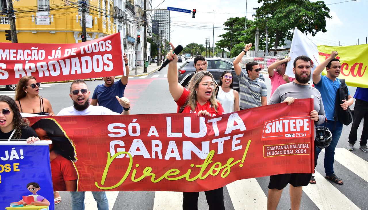 Com cartazes como "só a luta garante direitos" e "convoque os aprovados" diversas pessoas estão nas ruas do Recife gritando em manifestação. É possível ver uma mulher loira com as mãos erguidas com camiseta do sintepe, esta é Marília