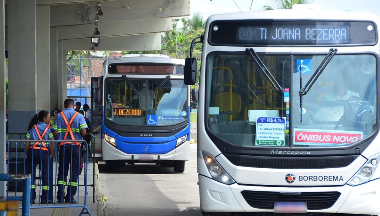 Imagem de um terminal integrado, é possível ver dois ônibus, um branco e um cinza com detalhes azuis. O branco está escrito "TI Joana Bezerra"
